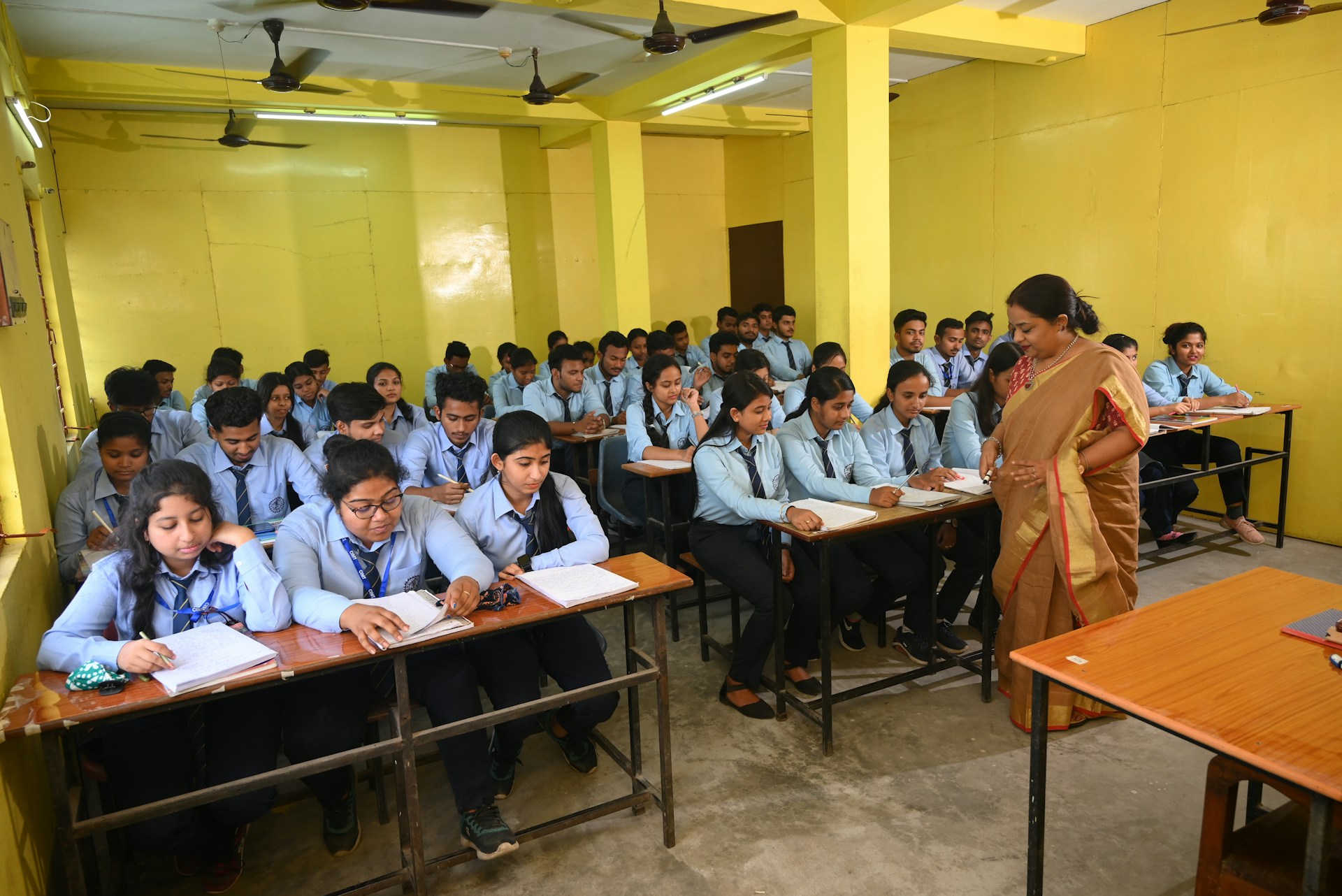 A classroom full of students in school uniforms sitting at desks with a teacher in a sari.