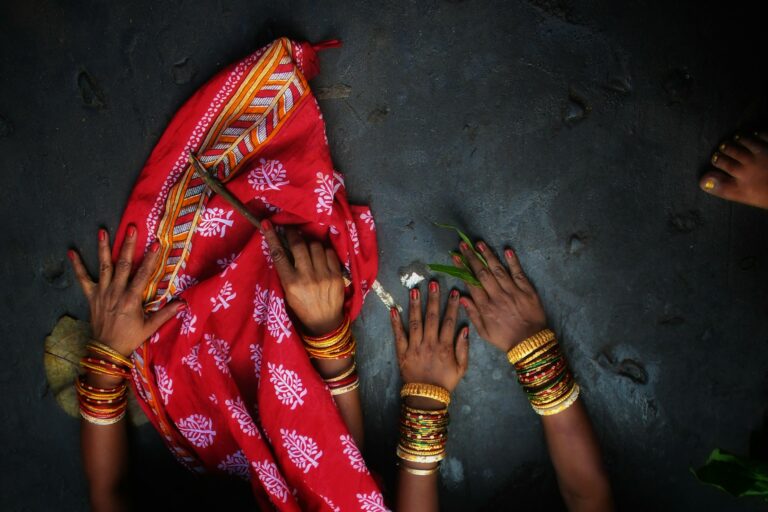 Multiple hands reaching toward a woman in red sari during a traditional ceremony, symbolizing community support and collective participation