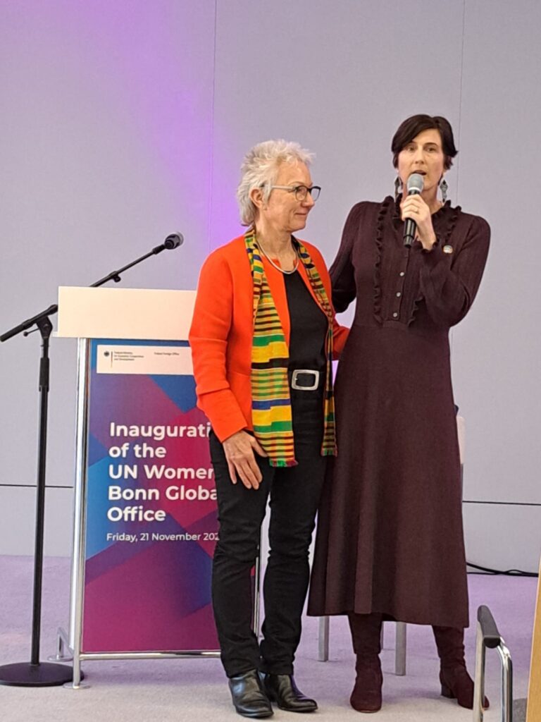 This photo captures a moment during the inauguration of the UN Women Bonn Global Office. Setting: The image is taken on a stage, in front of a backdrop displaying the event title, logos for the German Federal Ministry for Economic Cooperation and Development (BMZ) and the German Federal Foreign Office, and the date "Friday, 21 November 2025". A microphone stand is visible on the left. Individuals: Dr. Angela Langenkamp (left), Chairperson of UN Women Germany, is standing with her arm around her colleague. She is wearing a bright orange blazer over a black top, black trousers, a colourful striped scarf, and black ankle boots. Abigail Erikson (right), Head of the UN Women Office in Bonn, is speaking into a microphone. She is wearing a long, dark brown ruffled dress with button details and brown boots.