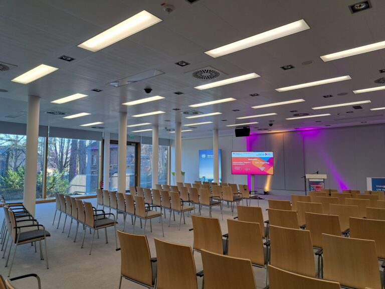 The image shows a modern conference room at the UN Campus in Bonn. The room is set up in a theater style with rows of wooden chairs. A stage is visible at the far end with a presentation screen and podium. The lighting is contemporary, with a mix of recessed and panel lights, and purple accent lighting on the back wall. Large windows along one side offer natural light and a view of the outdoors. The venue has been prepared for the inauguration of the UN Women Global Office.