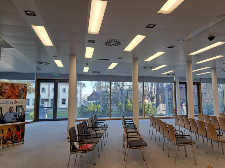 This is the Conference Room at the UN Campus in Bonn, which has floor-to-ceiling windows offering a view of the surrounding buildings and greenery. Rows of contemporary, empty chairs have been arranged for the inauguration of the UN Women Global Office Campus Bonn. Recessed and linear overhead lighting provides bright illumination. On the far left, an informational banner displaying the 'UN Women' logo is visible.