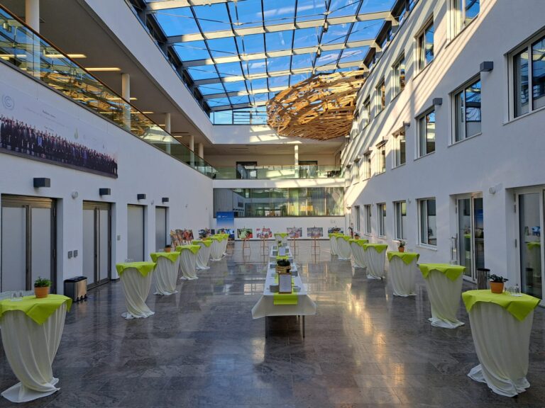 The image captures an event setup at the UN Campus in Bonn, Germany. The venue features a large, bright atrium with a glass roof and a distinctive, large, wooden art installation hanging from the ceiling, titled "Nest" by artist Tadashi Kawamata. Numerous cocktail tables draped in white cloths with lime green overlays are arranged in rows for the auguration of the UN Women Global Office in Bonn. A long table with a white tablecloth is set up in the center of the room.