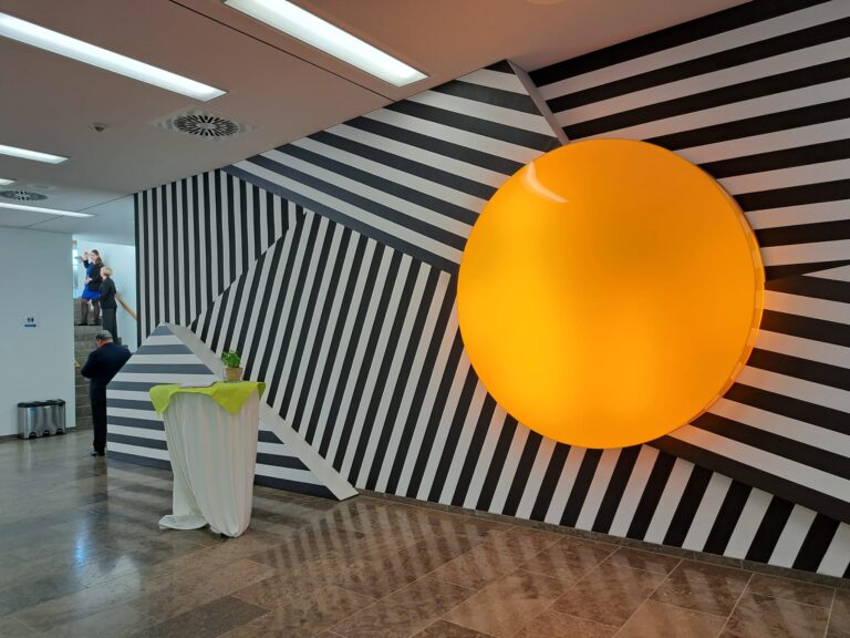 The image features the art installation "The Illuminator" by Wafae Ahalouch el Keriasti, a female artist from Morocco. The installation is located in the former German parliamentary building in Bonn, which now serves the UN Campus. The work is a large, illuminated orange sphere set against a backdrop of black-and-white stripes. The sphere represents the sun. The artist used the contrast of black and white stripes with the orange sphere to symbolize global warming.